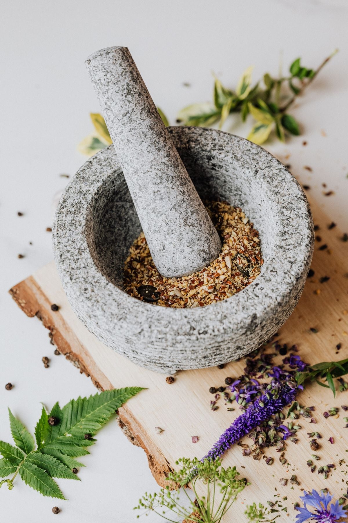 A granite mortar and pestle of milled seed on a pale stone surface, botanicals alongside